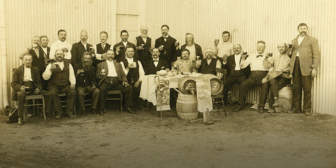 An old-timey black-and-white photo of around 20 people toasting with Belgian beer glasses