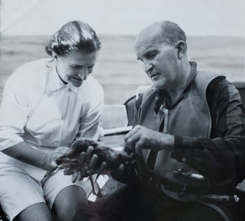 Two people in a small boat, one of them wearing diving gear, peer intently at an artifact that the diver is holding