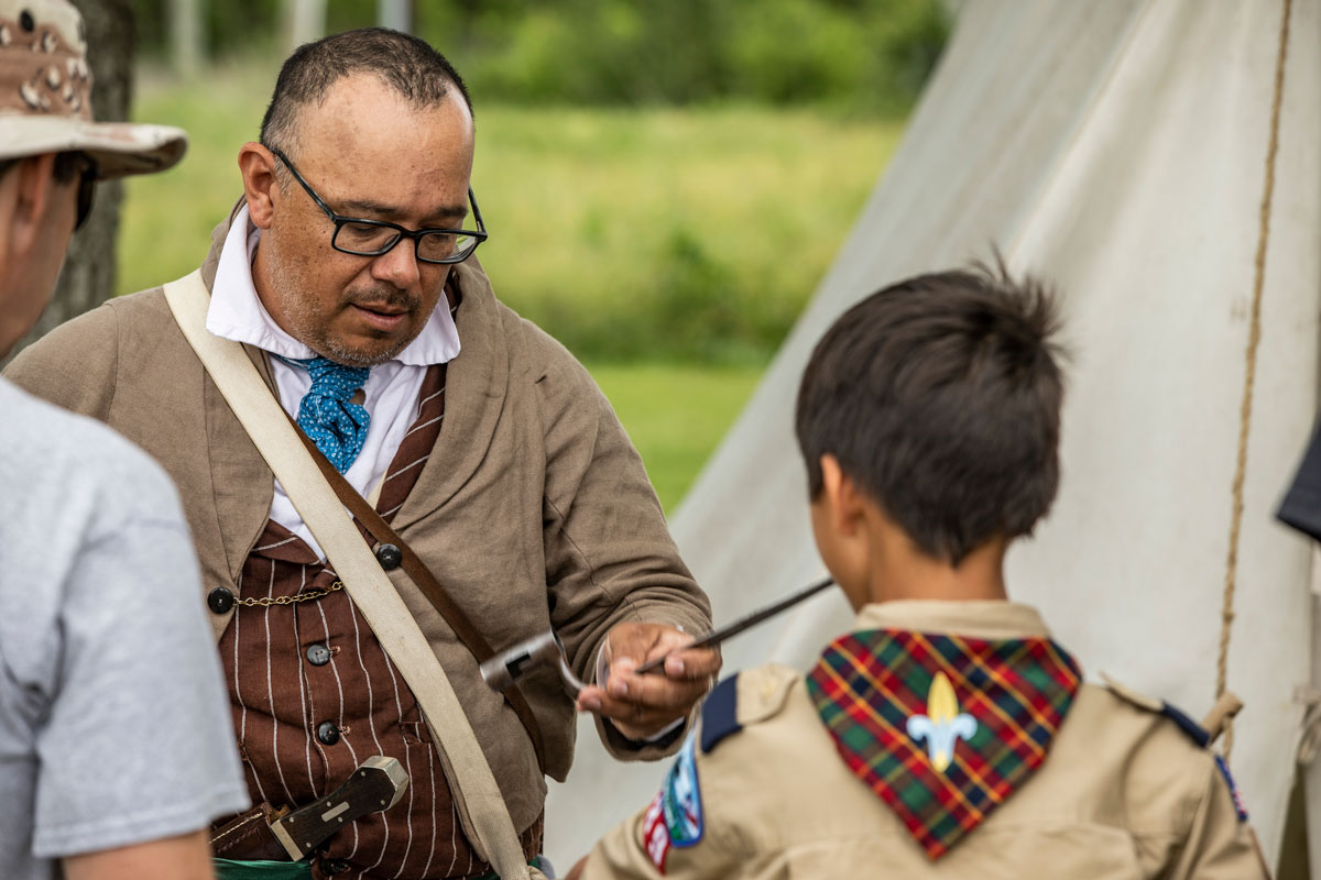 A historical reenactor in 1800s period clothing speaks to a young boy in a Boy Scout uniform.