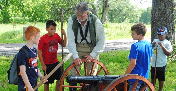 a man dressed in a historic outfit showing 4 boys how to load a cannon