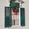 A green Christmas wreath with a red bow hanging above the window of a white building