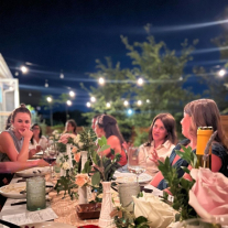 A group of people gather around a full dinner table under patio lights at twilight. 