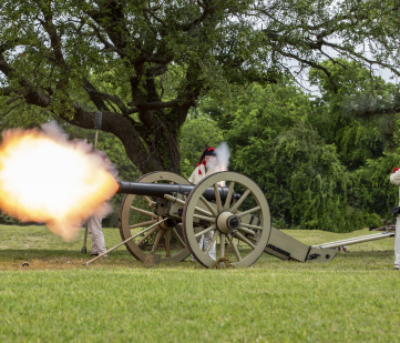 A cannon being fired. Two men in historic outfits stand near the cannon. A plume of fire is coming out the end of the cannon.