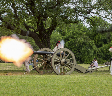 A cannon being fired. Two men in historic outfits stand near the cannon. A plume of fire is coming out the end of the cannon.